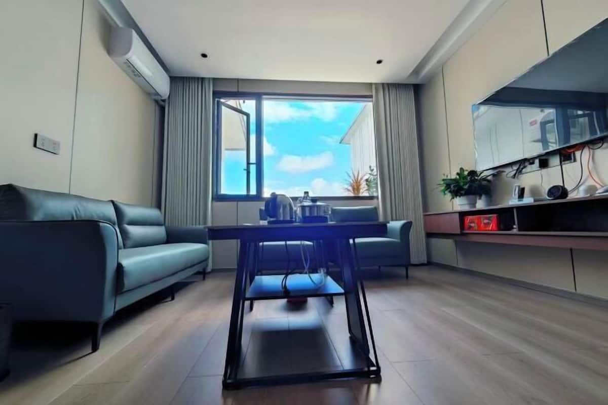 Ein modernes Wohnzimmer mit einer Couch, einem Esstisch, einem an der Wand befestigten Fernseher und einem großen Fenster mit Vorhängen.