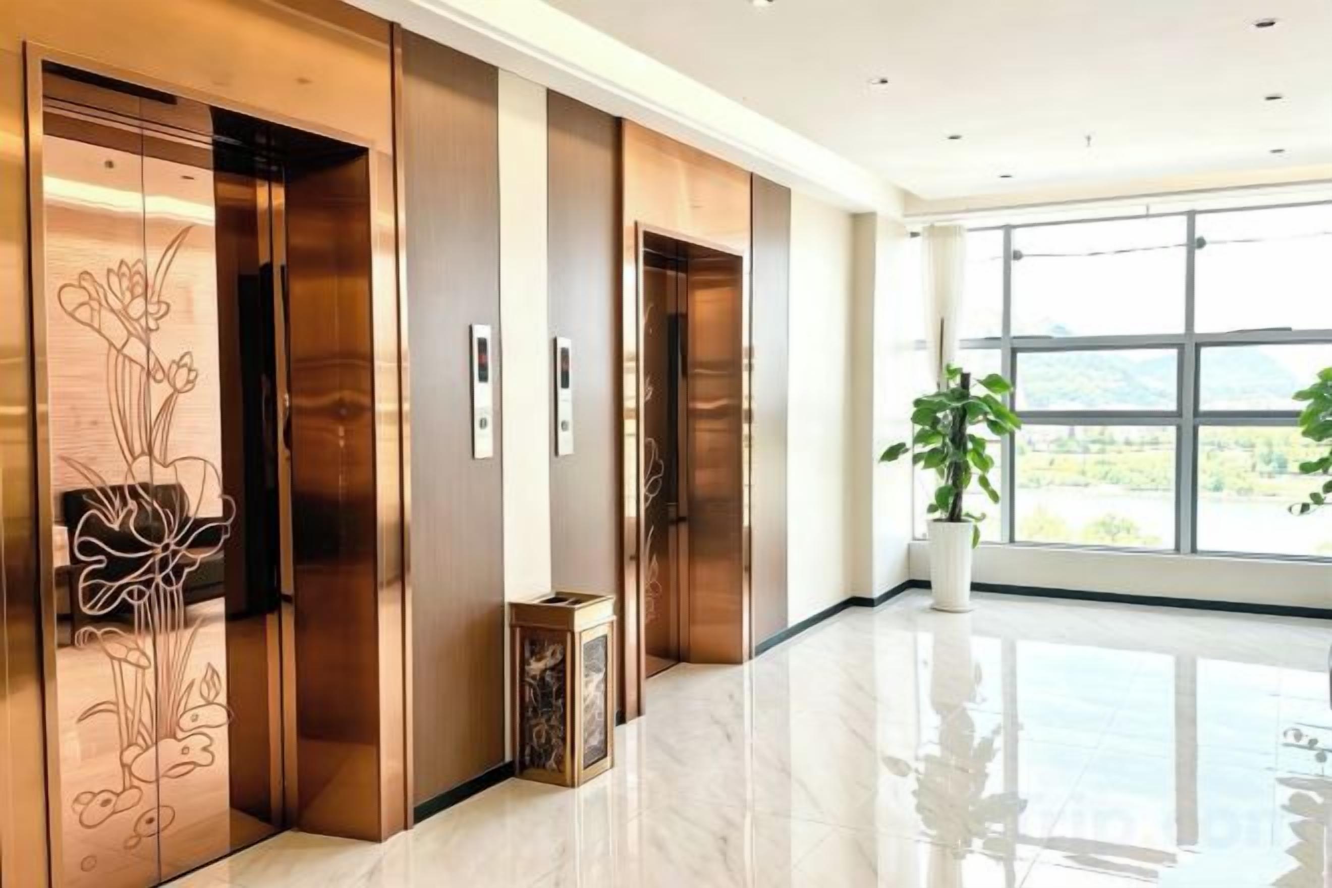 A modern hotel lobby with a polished floor, wooden elevator doors with floral designs, and a large window offering a view of the outdoors.
