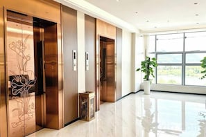 A modern hotel lobby with a polished floor, wooden elevator doors with floral designs, and a large window offering a view of the outdoors. - Hongfurong Hotel (Anlong)