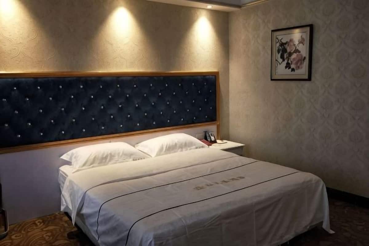 Ein Hotelzimmer mit einem gepolsterten Kopfteil, einem ordentlich bezogenen Bett mit weißer Bettwäsche, einem Nachttisch mit Lampe sowie einem gerahmten Bild an der Wand.