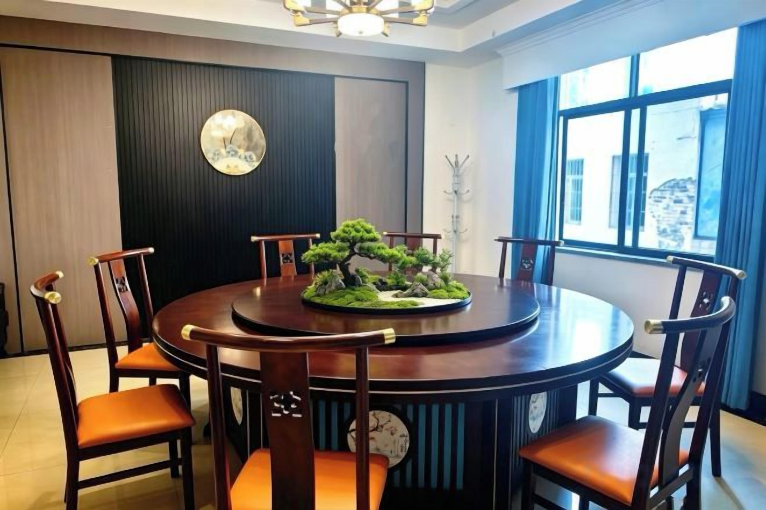 A round dining table with six wooden chairs, a bonsai tree centerpiece, and a wall-mounted clock.