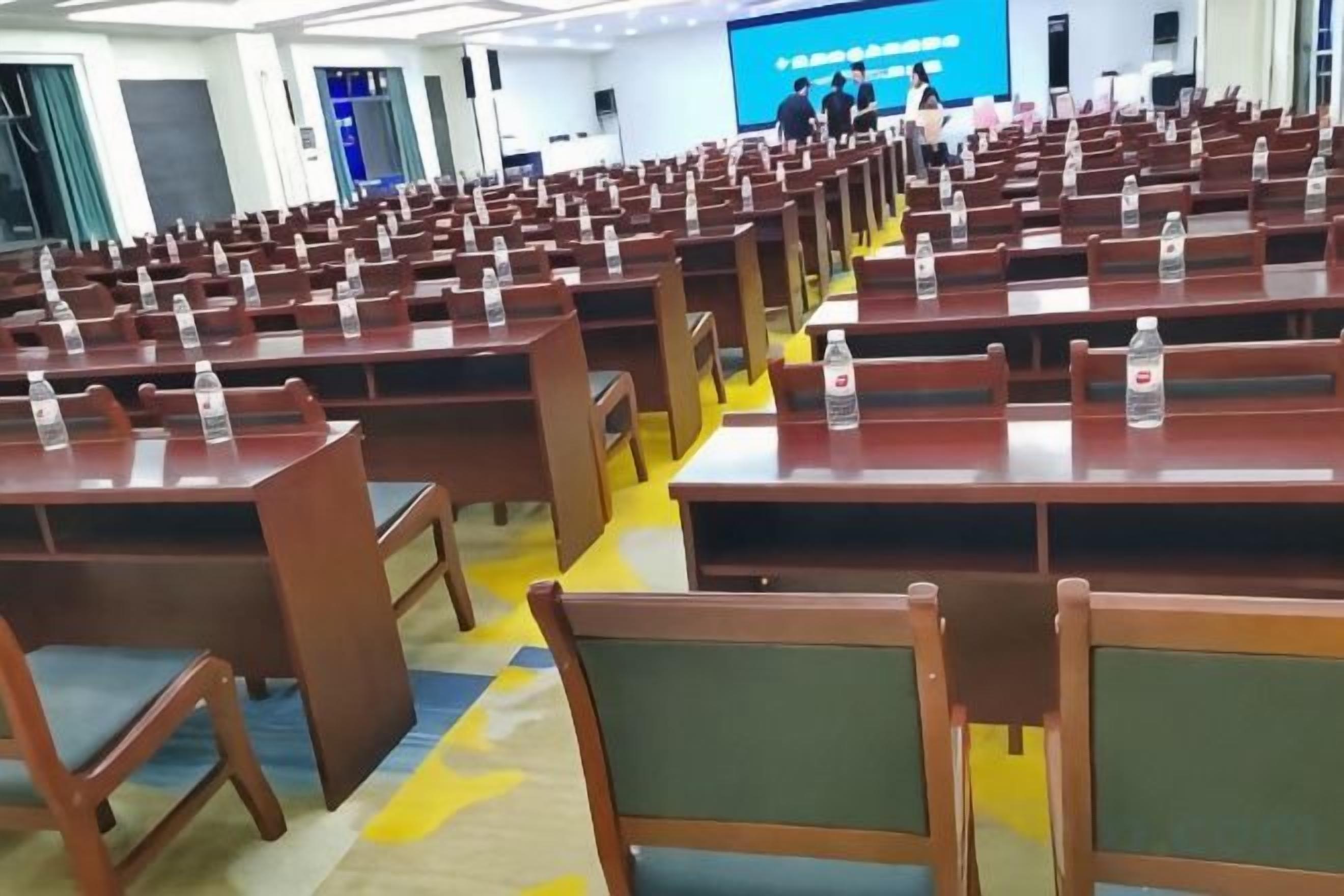 Un salón de conferencias con filas de mesas y sillas de madera, algunas con botellas de agua encima.