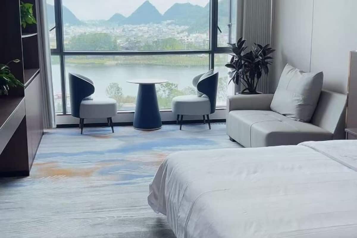 Ein Hotelzimmer mit einem Bett, einer Couch, zwei Sesseln, einem kleinen Tisch und einem großen Fenster mit Blick auf einen See und Berge.