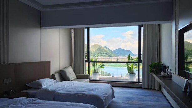 Habitación de hotel con dos camas, un ventanal con vistas a montañas y un cuerpo de agua, y un balcón con plantas.