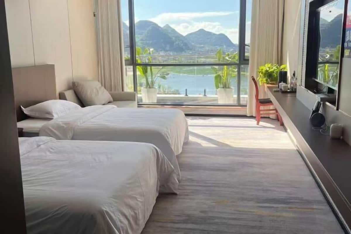 Ein Hotelzimmer mit zwei Betten, einem Fernseher, einem Schreibtisch mit Stuhl und einem Balkon mit Blick auf Berge und Wasser.