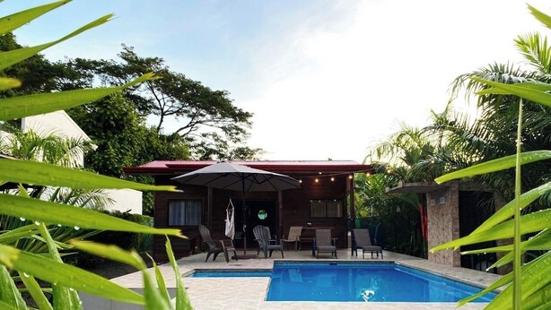 Una piscina con un edificio de techo rojo, mobiliario exterior y abundante vegetación.