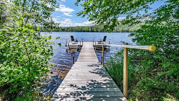 En träbrygga som leder ut till en sjö, med två stolar och ett bord, omgiven av grönska.