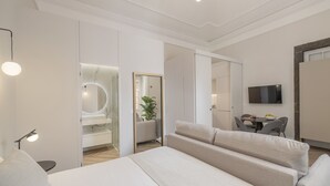 A modern hotel room with a large bed, a bedside table with a lamp, a seating area with a sofa and chairs, a small table with a bowl of fruit, and a TV mounted on the wall. - Luxury, art and elegance - FAA Belas Artes 2 (Funchal)
