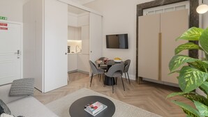A modern living room with a sofa, a coffee table, a dining area with chairs, a TV mounted on the wall, and a large plant. - Luxury, art and elegance - FAA Belas Artes 2 (Funchal)