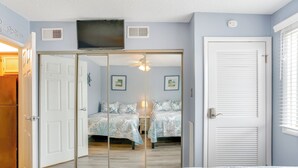 Une chambre avec un placard à portes coulissantes, un lit recouvert d’une couvre-lit à motifs, un ventilateur de plafond et un téléviseur fixé au mur.