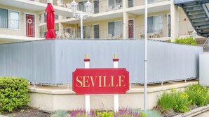 Un panneau rouge avec le mot «SEVILLE» écrit en lettres dorées, entouré d’un jardin où poussent différentes plantes et surmonté d’un parasol rouge.