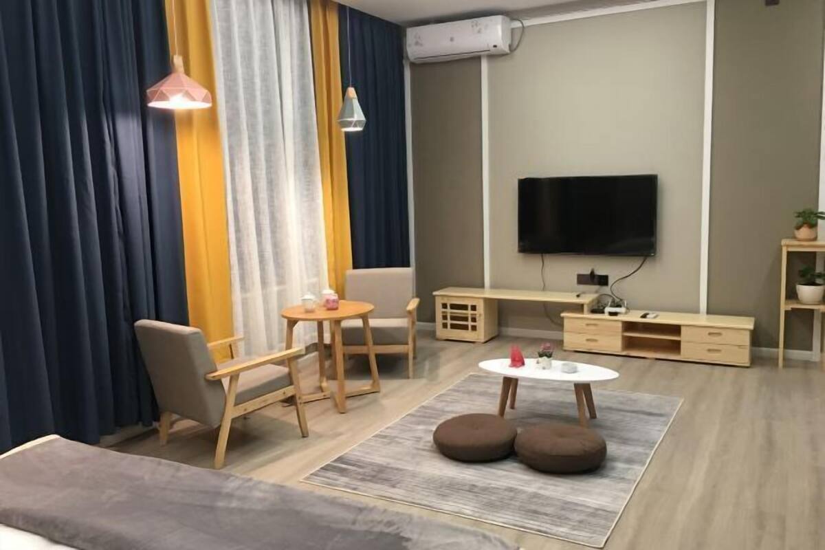 A modern living room with a flat-screen TV, wooden furniture, and a large window with curtains.