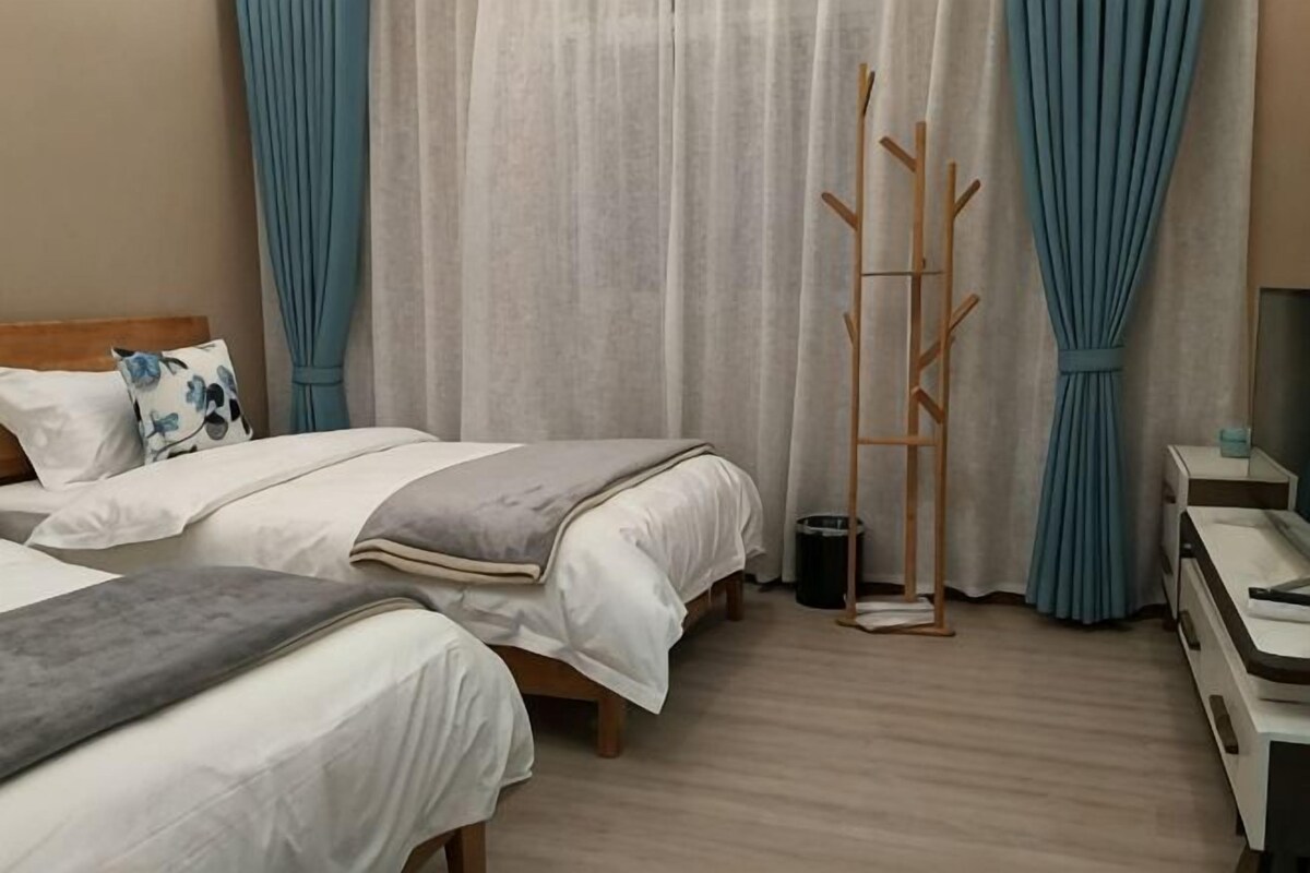 A hotel room with two beds, a bedside table, a mirror, a wooden coat rack, and a window with curtains.