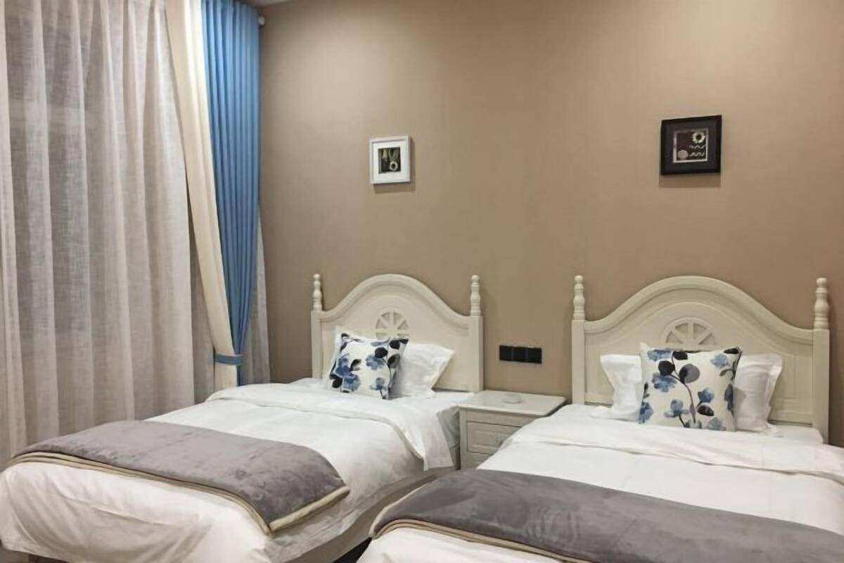 A hotel room with two beds, a nightstand, a wall clock, and a window with curtains.