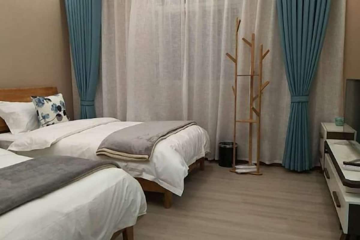 A hotel room with two beds, a bedside table, a mirror, a wooden coat rack, and a window with curtains.
