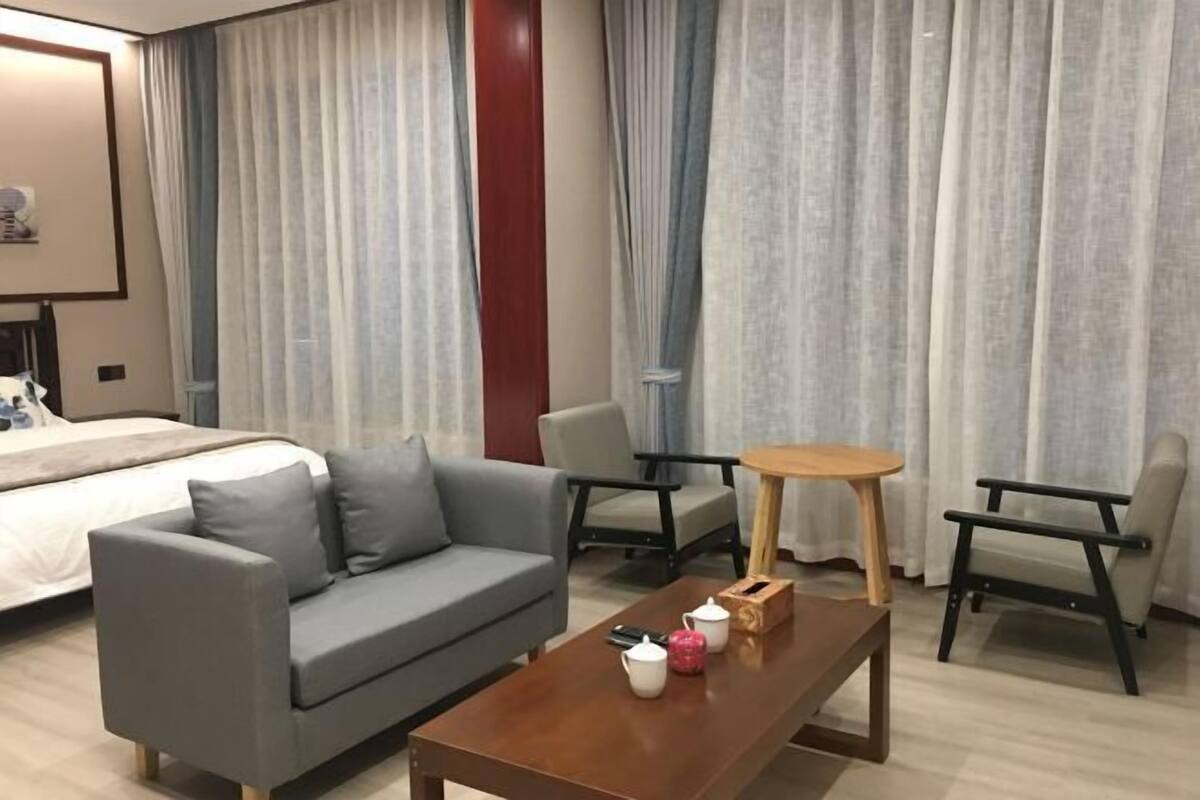 A hotel room with a bed, grey sofa, wooden coffee table, two chairs, and a small table with a vase and teapot.