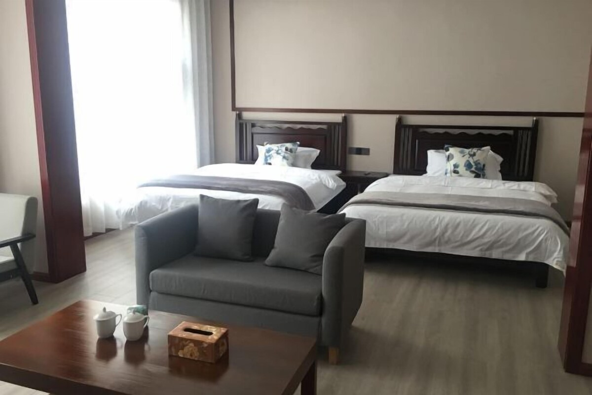 A hotel room with two beds, a grey sofa, a wooden coffee table with a tissue box, and a small table with a teapot and cup.