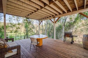 An outdoor wooden deck with a round table and chairs, a grill, and a view of a natural landscape.