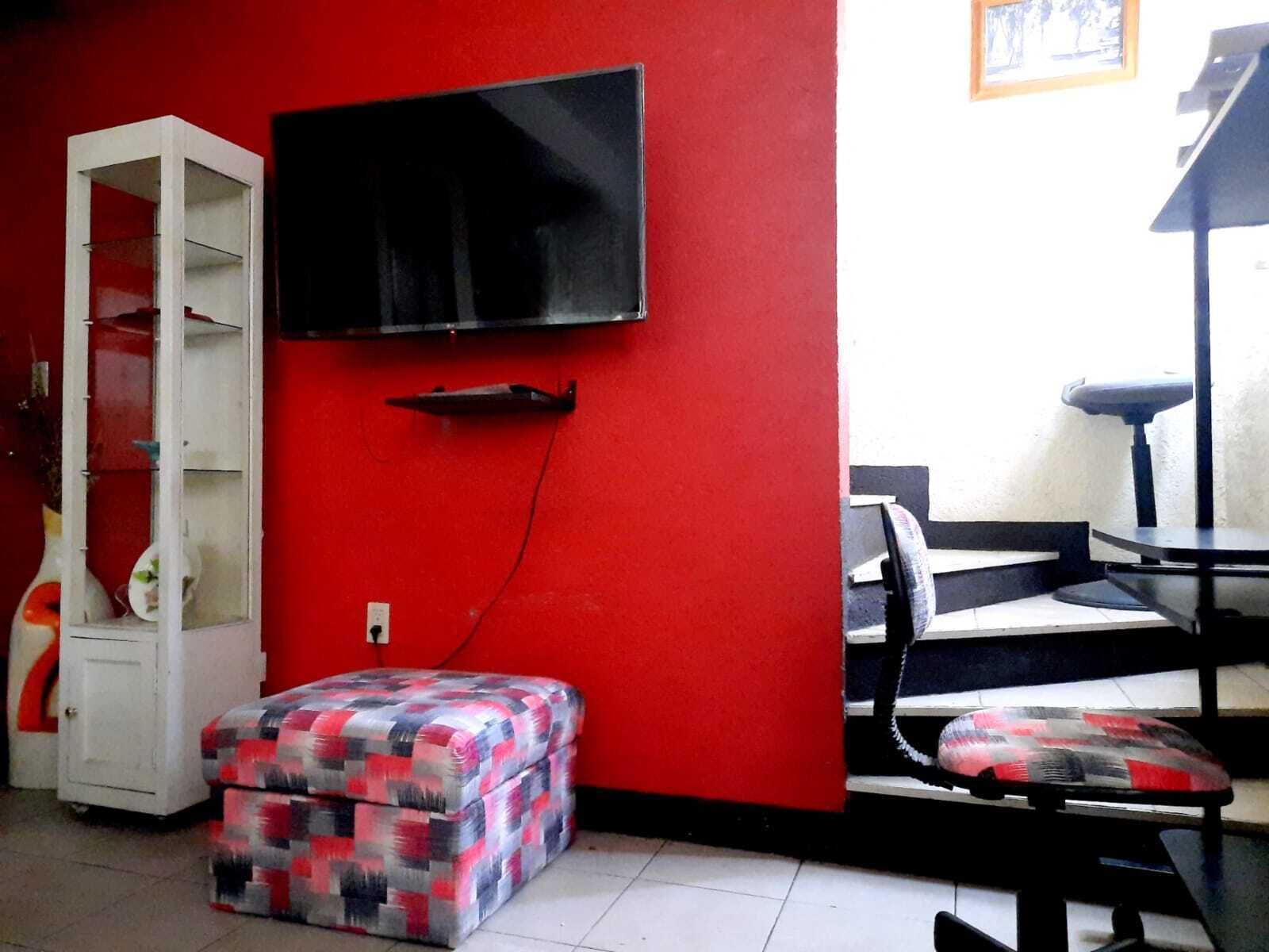 Quarto com parede vermelha, armário branco, televisão presa na parede, banqueta estampada e cadeira preta e branca.