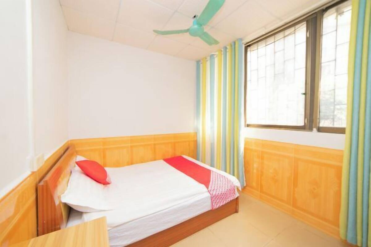 A single bed with a wooden headboard, a ceiling fan, and a window with striped curtains.
