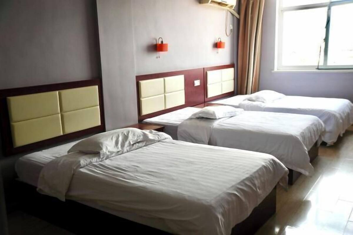 A hotel room with three single beds, a wooden headboard, a window with curtains, and a wall-mounted light fixture.