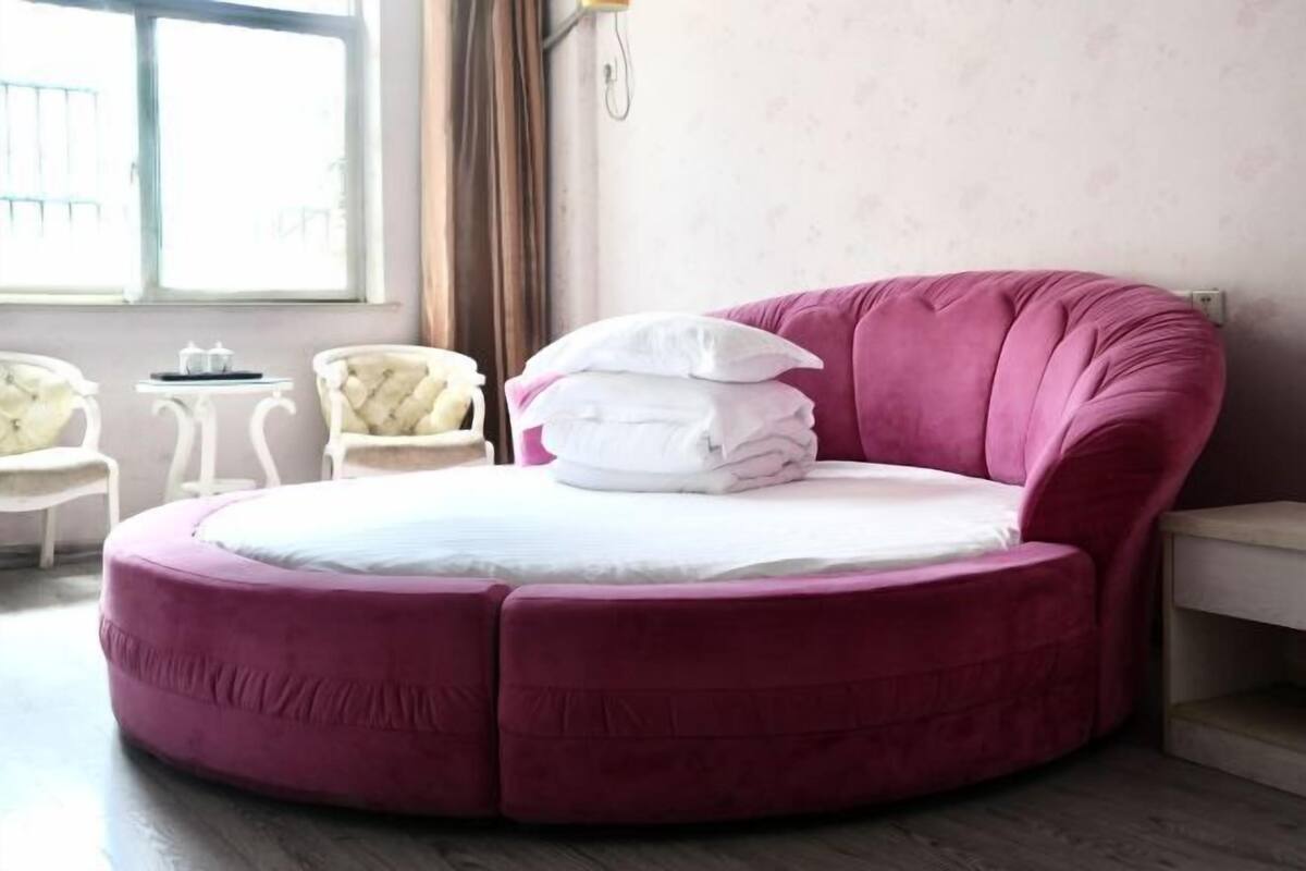 A circular bed with a pink upholstered headboard, white pillows, and a white mattress. There are two small white tables with chairs, a window with curtains, and a wall-mounted light fixture.