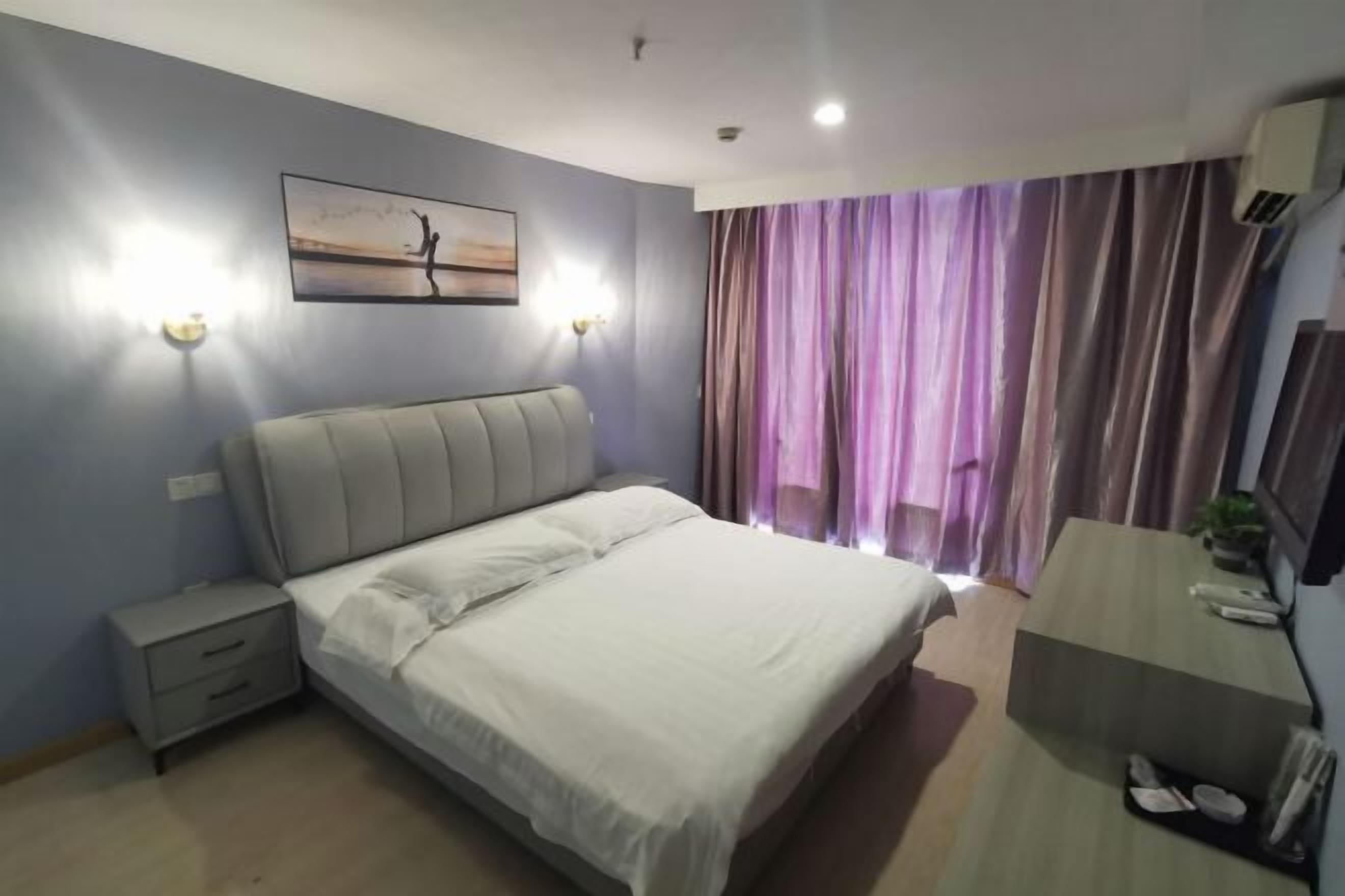 A bedroom with a bed, bedside tables, a nightstand, a desk, a chair, a window with curtains, and a wall-mounted air conditioner.