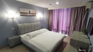 A bedroom with a bed, bedside tables, a nightstand, a desk, a chair, a window with curtains, and a wall-mounted air conditioner.
