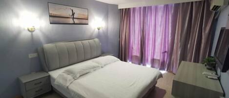 A bedroom with a bed, bedside tables, a nightstand, a desk, a chair, a window with curtains, and a wall-mounted air conditioner.