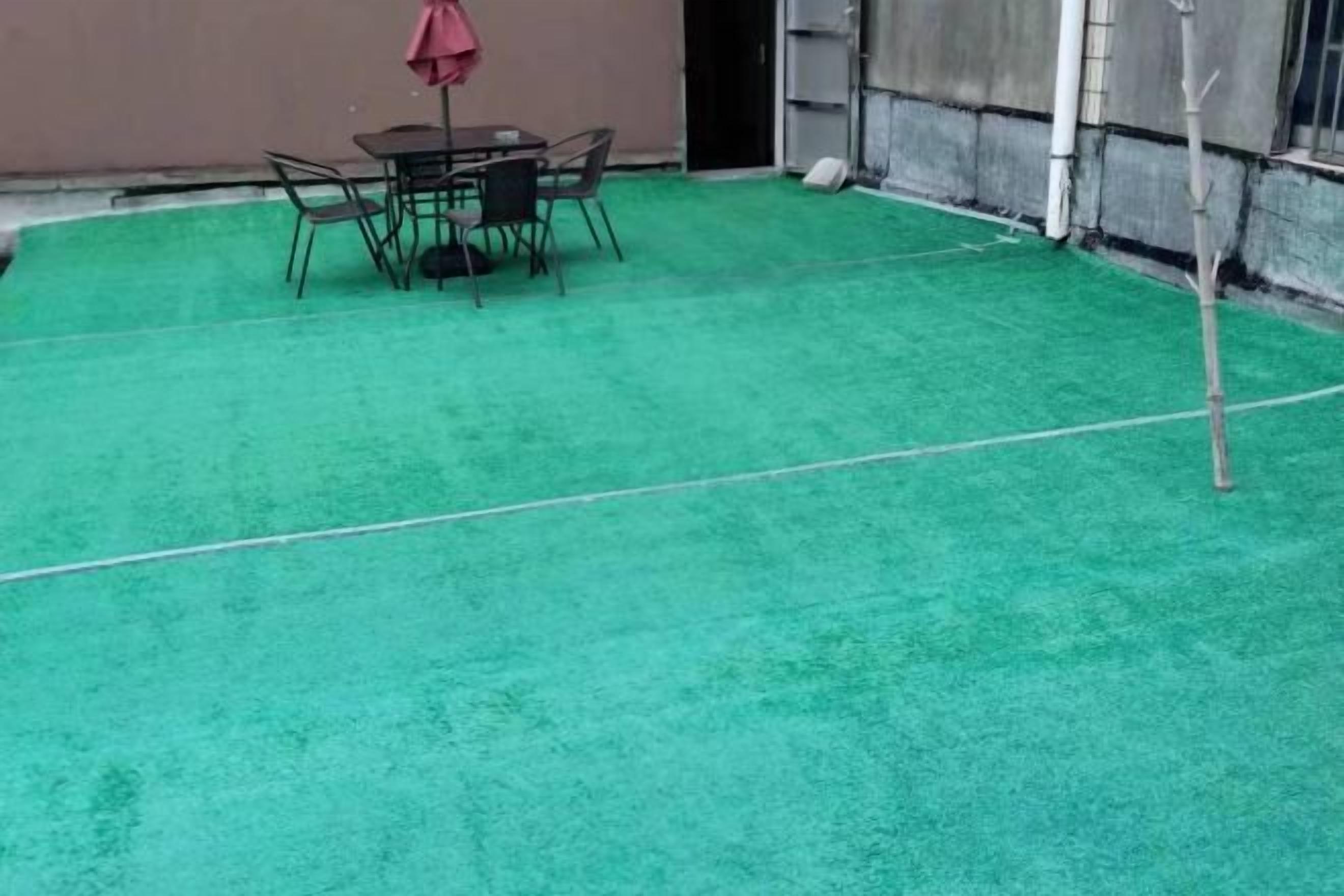 Un espace recouvert d’une pelouse artificielle verte, avec une table et des chaises, un parasol rouge et un petit arbre.