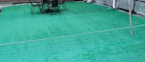 A green artificial turf area with a table and chairs, a red umbrella, and a small tree.