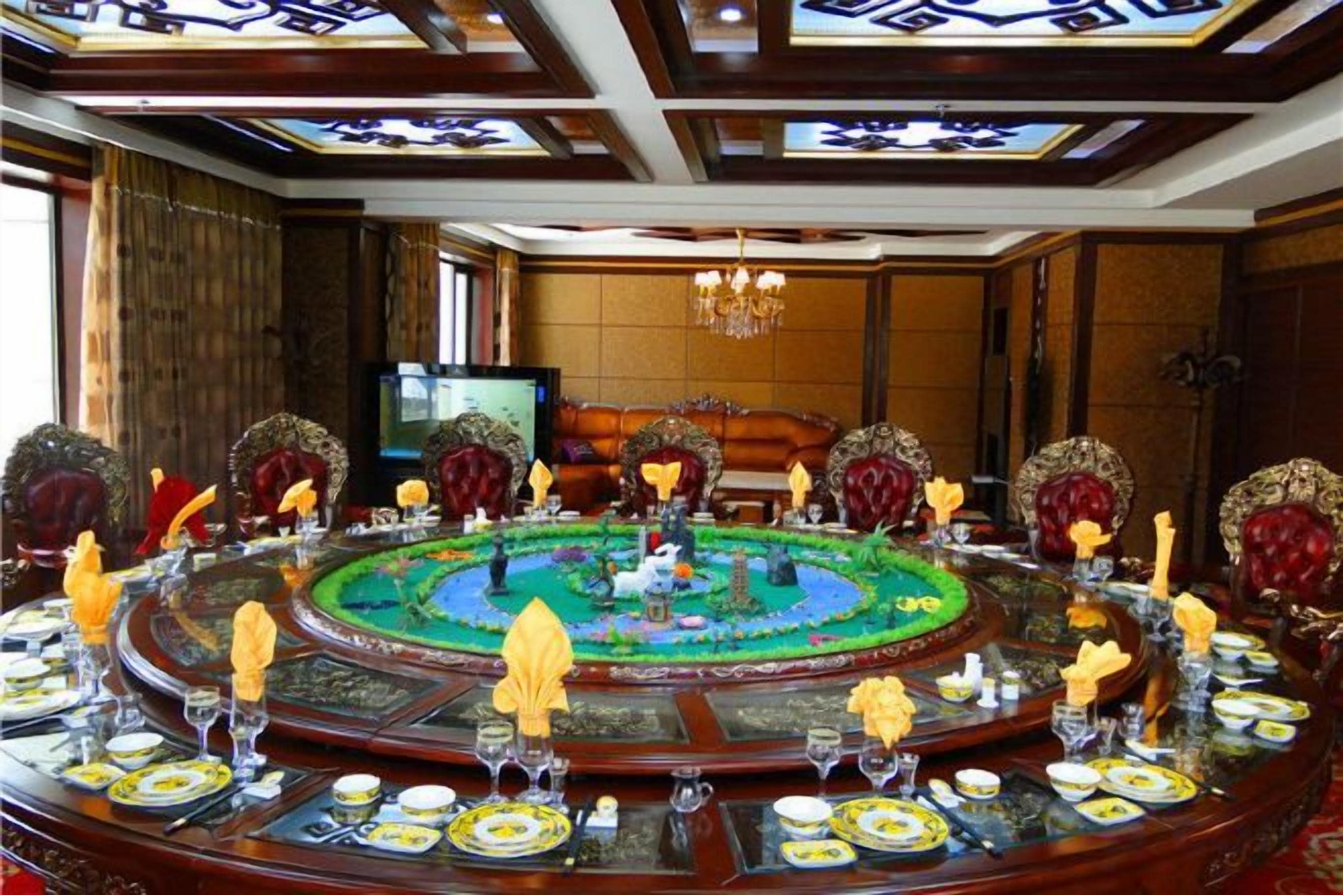 A round table with a central decorative piece, surrounded by chairs with ornate backrests, in a room with a chandelier and a television in the background.