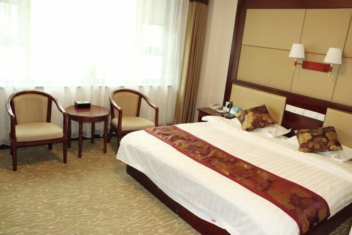 Ein Hotelzimmer mit einem großen Bett, zwei Sesseln, einem kleinen Tisch, einem Nachttisch und einem Fenster mit Vorhängen.