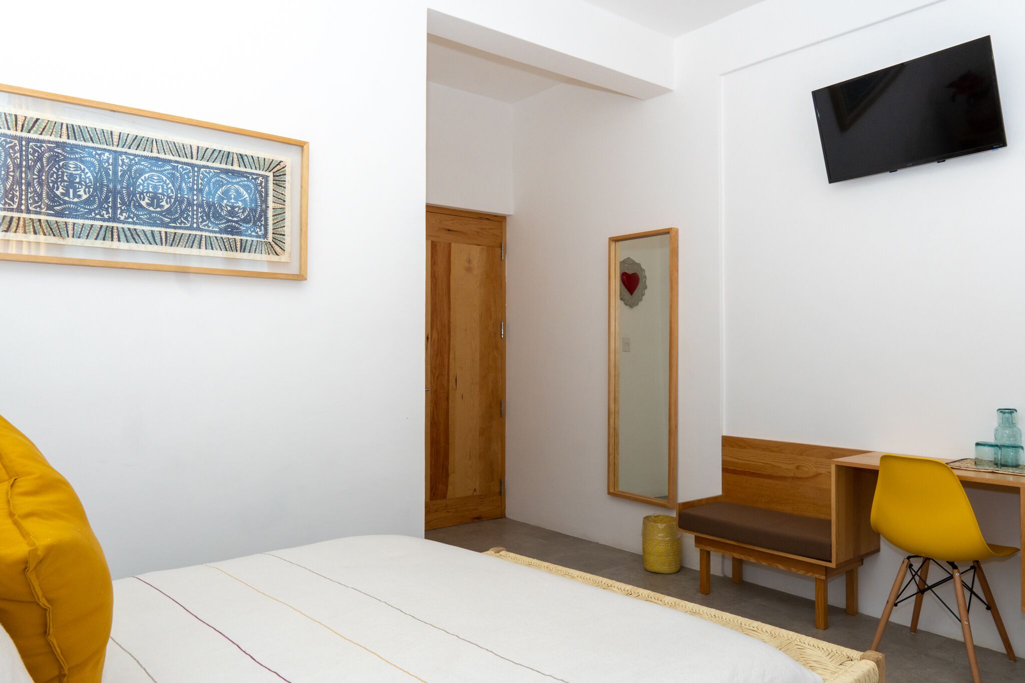 A bedroom with a bed, a desk, a chair, a TV, a mirror, and a framed artwork.