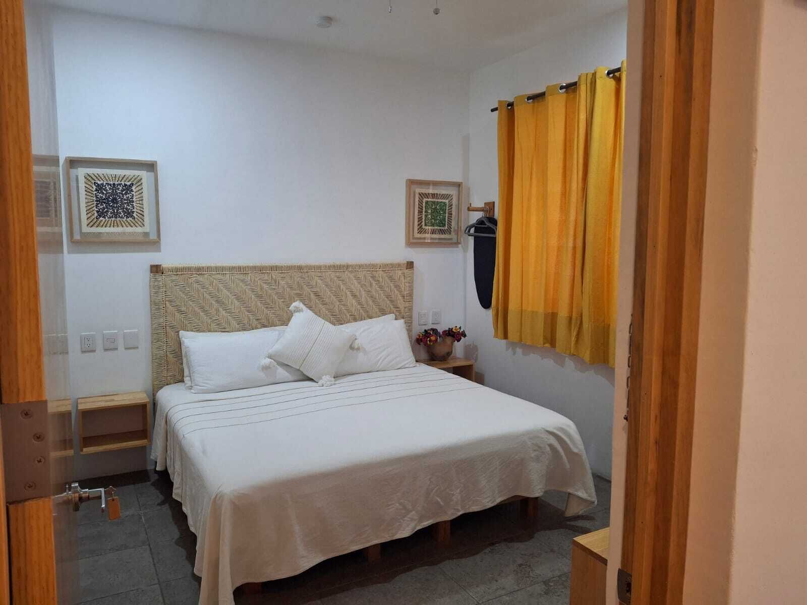 A bedroom with a bed, a nightstand, a wall-mounted shelf, and a window with curtains.