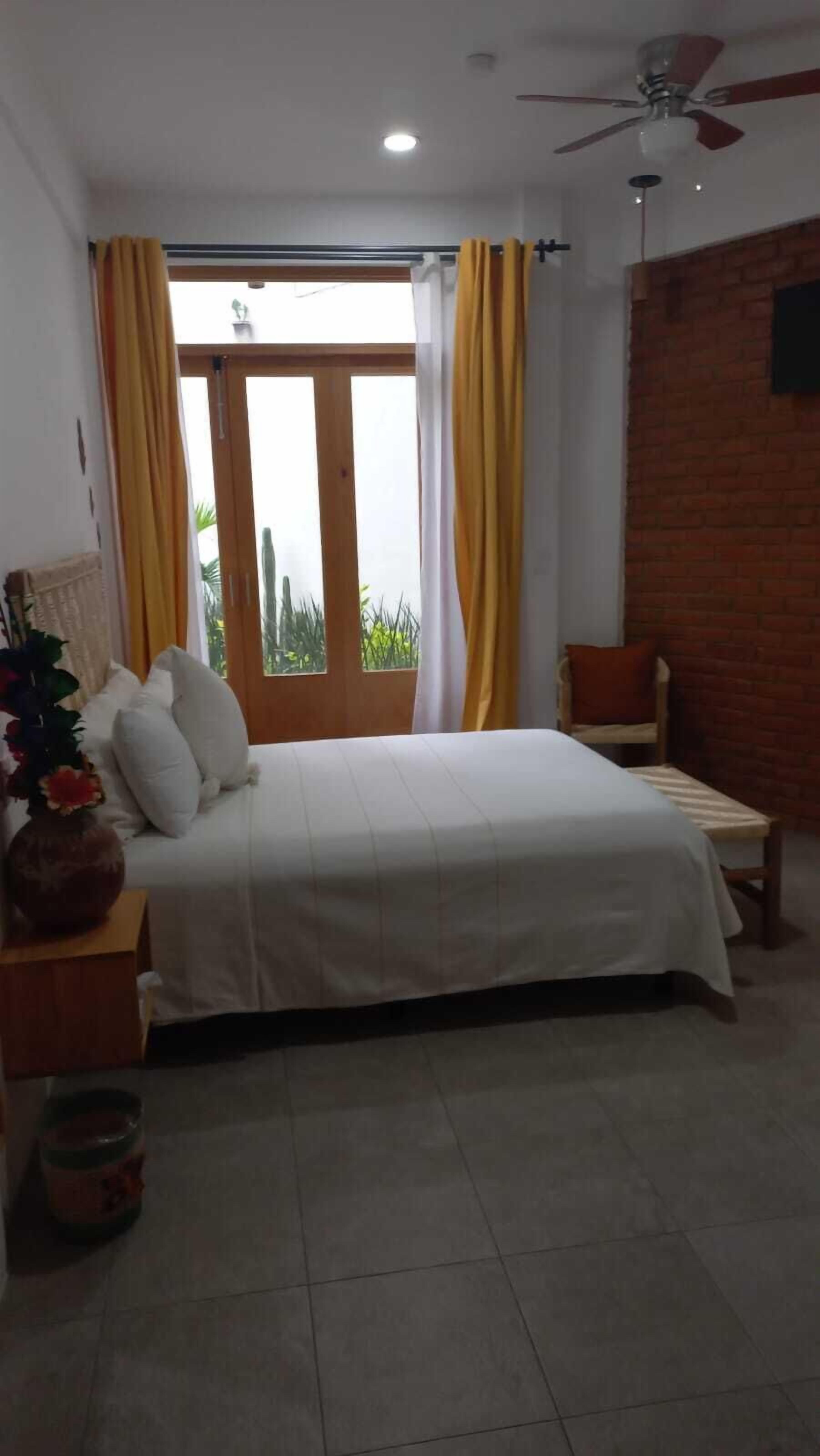 A bedroom with a bed, a nightstand with a vase, a window with curtains, and a brick wall.