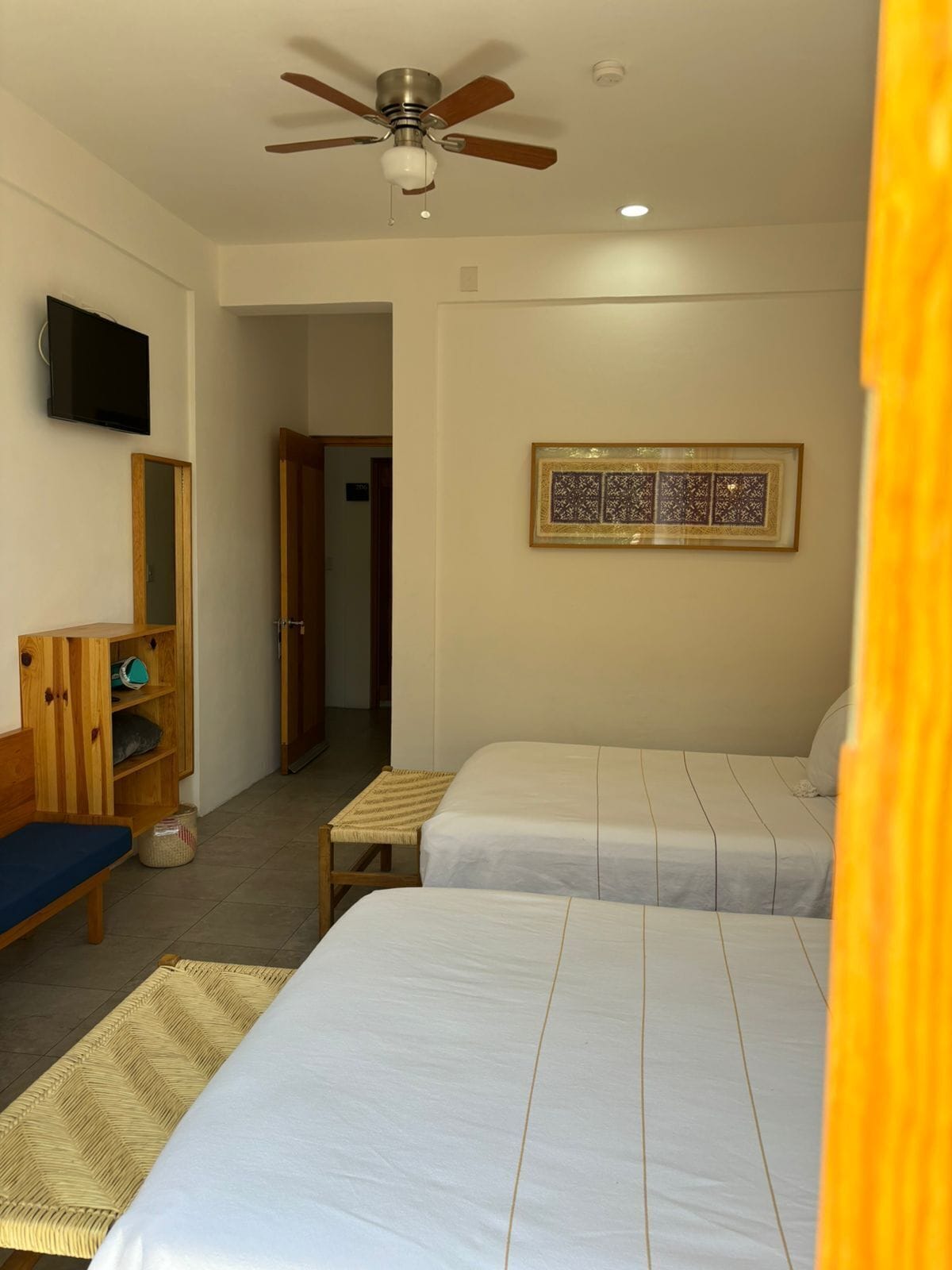 A hotel room with two beds, a ceiling fan, a television, a wooden cabinet, and a framed artwork on the wall.