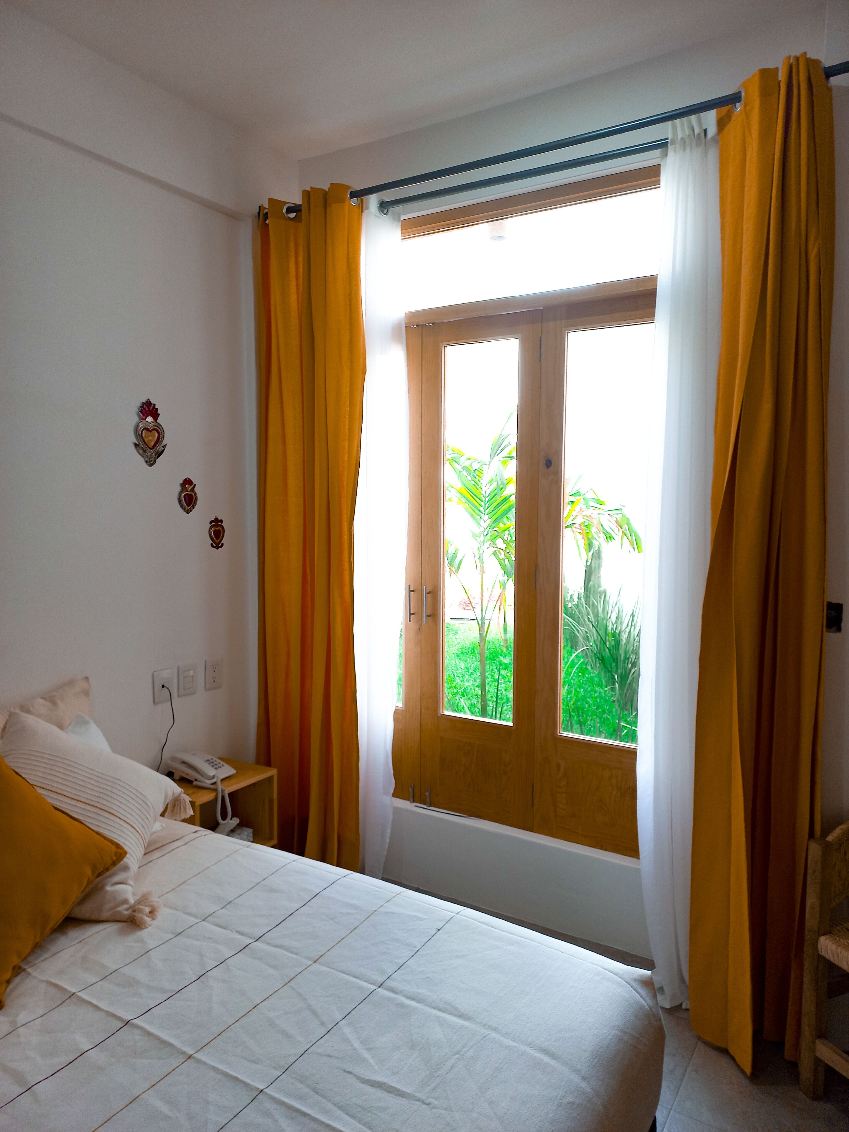 Une chambre à coucher avec un lit, une table de chevet sur laquelle se trouve un téléphone, une fenêtre avec des rideaux et une vue sur la verdure à l’extérieur.