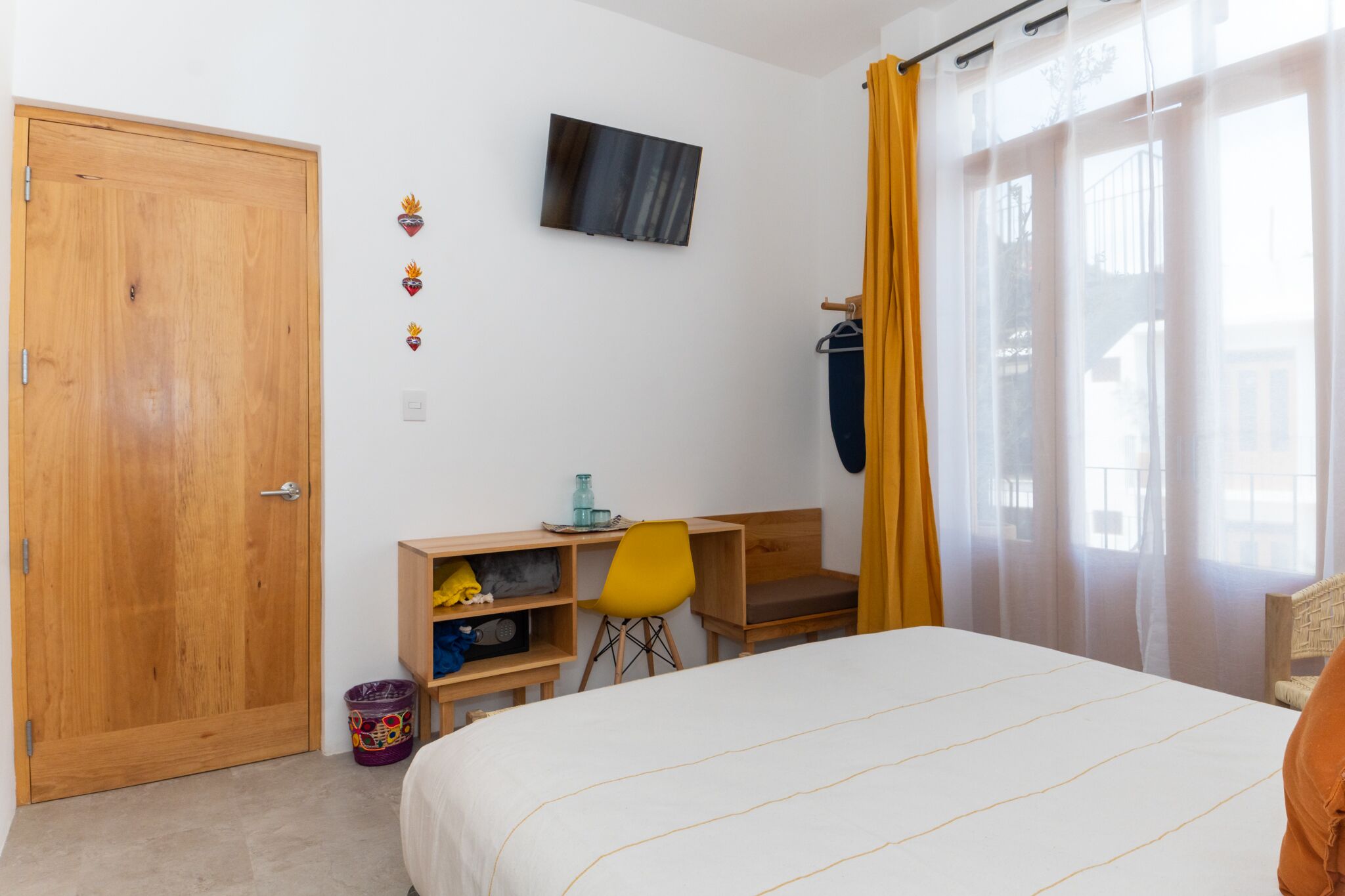 A bedroom with a bed, a desk with a chair, a TV on the wall, and a window with curtains.