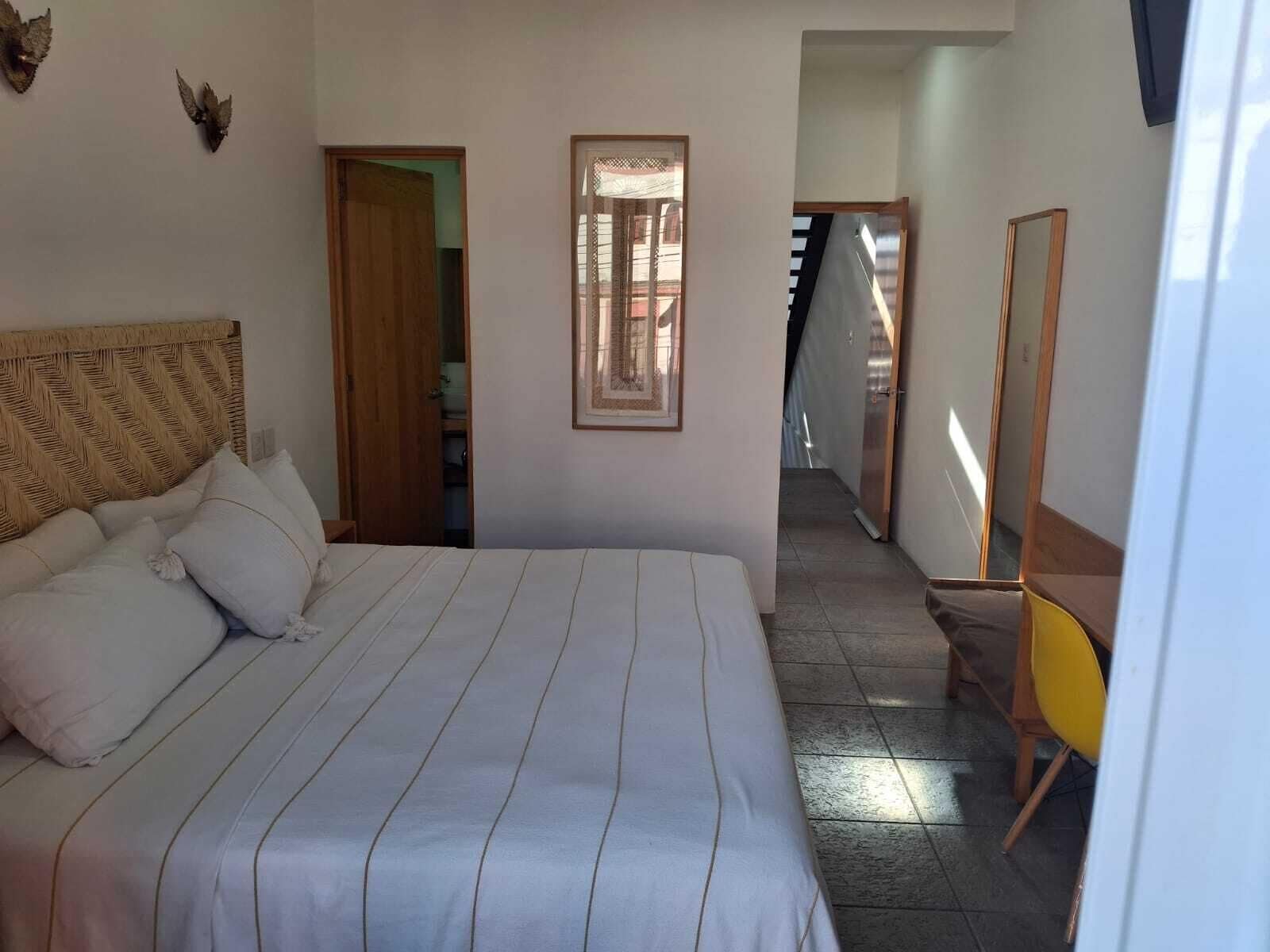Habitación de hotel con una cama grande, cabecera de madera, un cuadro enmarcado en la pared, una silla amarilla y un espejo.