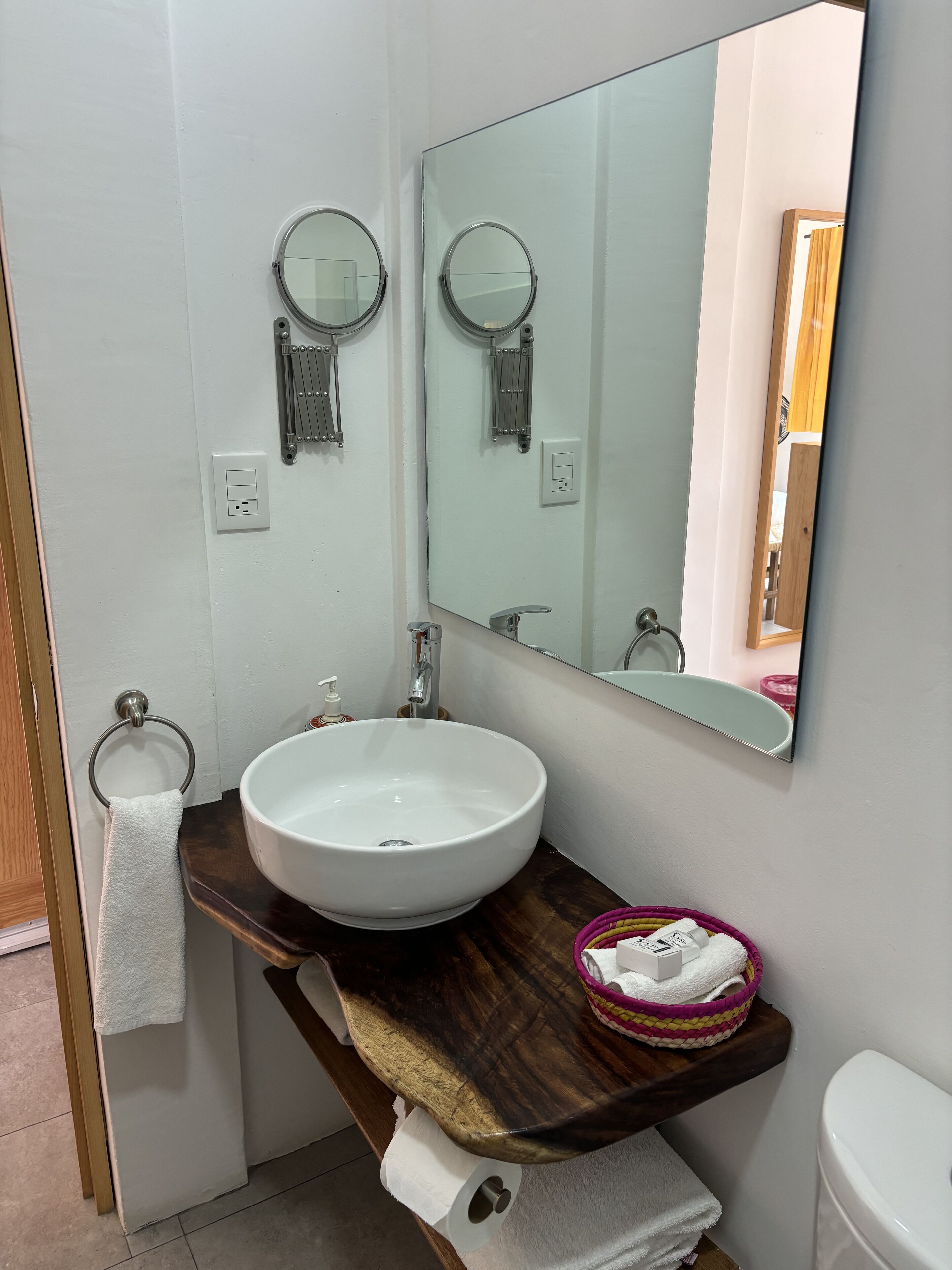 Un baño con lavamanos blanco, un espejo grande, un perchero para toallas y un mueble de tocador de madera.