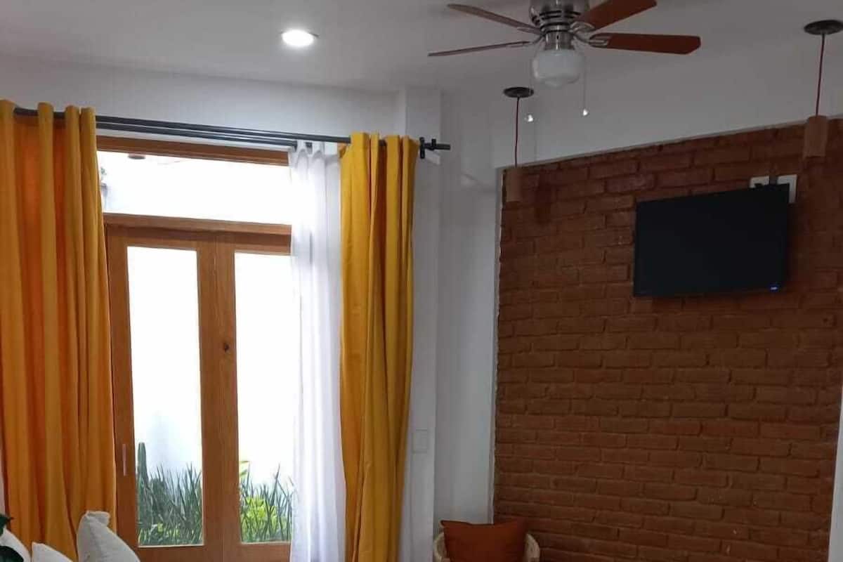 A bedroom with a bed, a chair, a TV mounted on a brick wall, and a ceiling fan.