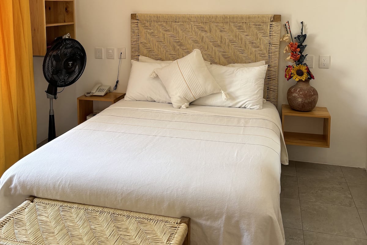 A neatly arranged bedroom with a bed, bedside table, fan, and a small wooden shelf.