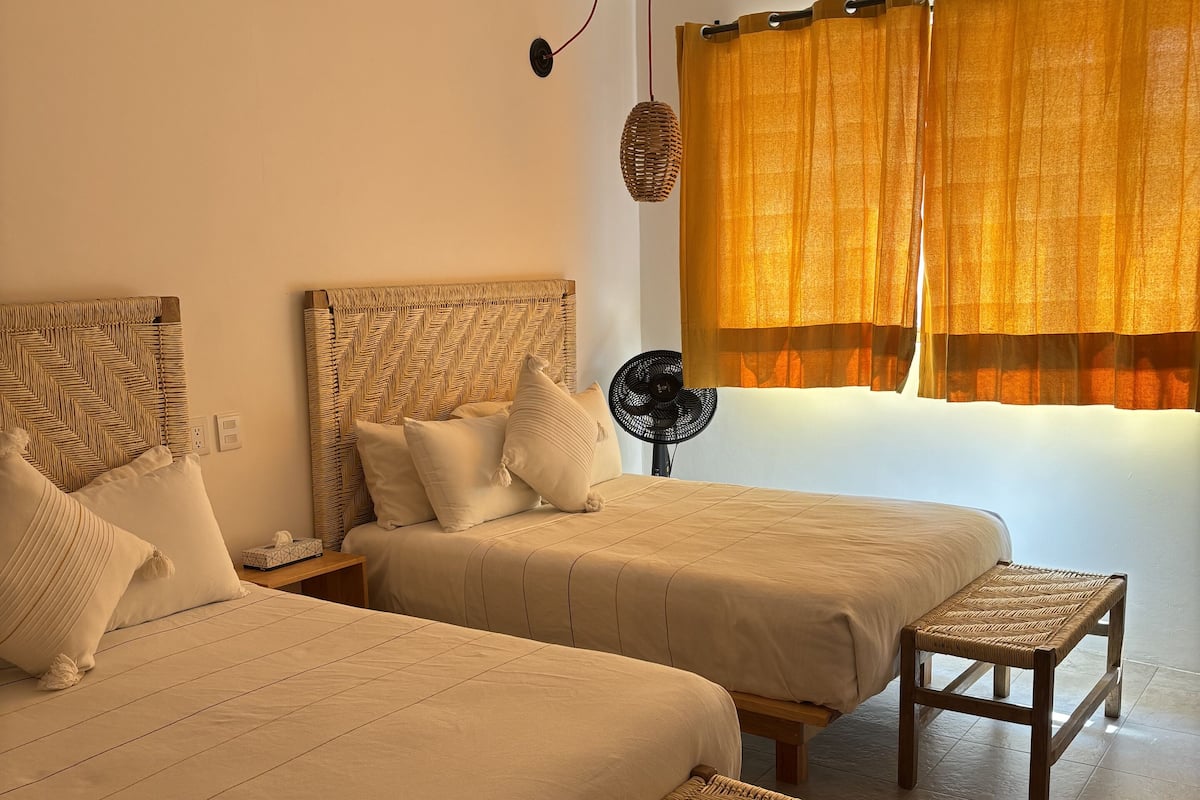 A room with two beds, a small table, a fan, and a window with curtains.