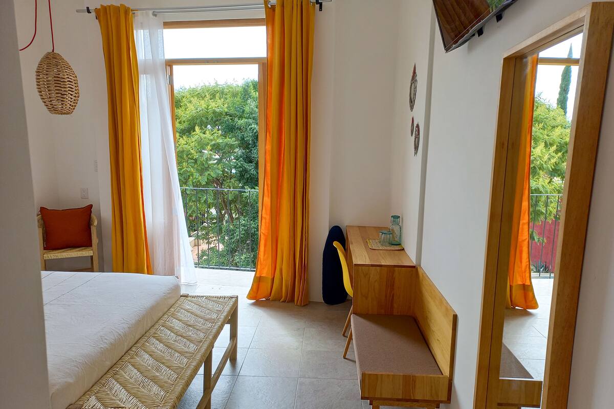 A room with a bed, a bench, a desk, and a chair. There is a window with curtains, a balcony, and a view of greenery outside.