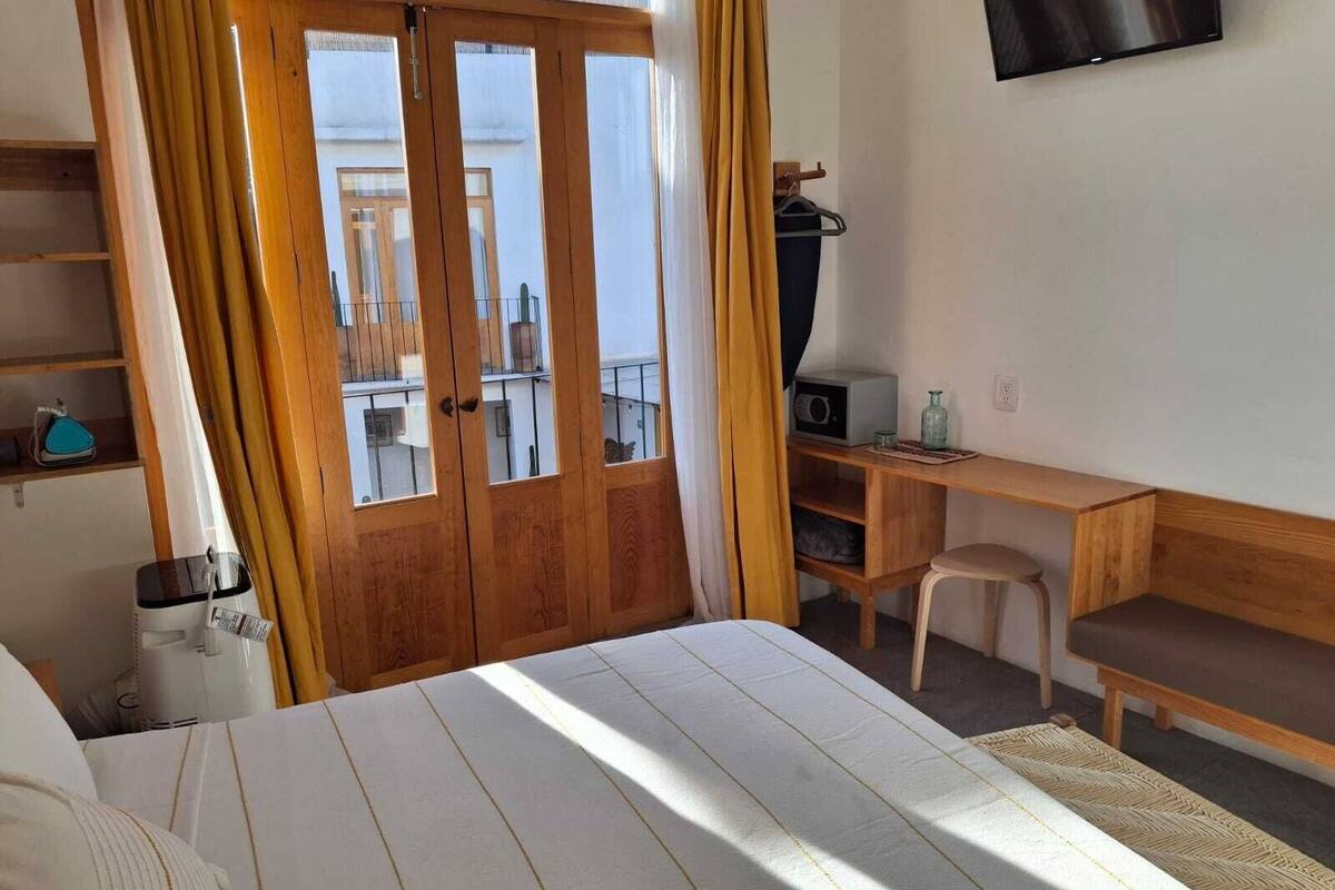 A bedroom with a bed, a wooden desk, a chair, a TV, and a balcony with curtains.