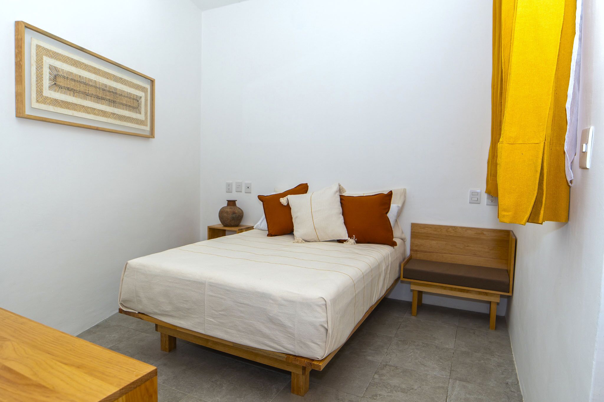A bedroom with a bed, a wooden bench, a small table, and a wall-mounted shelf.