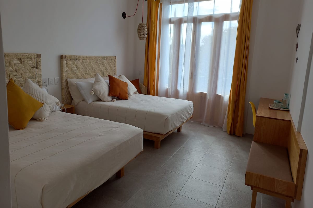 A hotel room with two beds, a wooden desk, and a large window with yellow curtains.