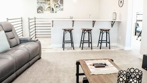 Een moderne woonkamer met een grijze bank, een houten salontafel en drie zwarte krukken. Achtergrond toont een keuken met een wit aanrechtblad en hangende吊り下げ灯.