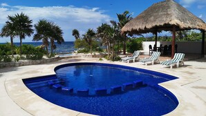 Una piscina infinita curvada con azulejos azules, rodeada de sillones de descanso y una estructura con techo de paja.